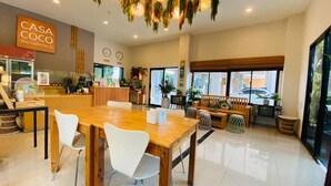 A modern hotel lobby with a wooden reception desk, white chairs, a wooden dining table, and a hanging plant chandelier. - Casa Coco Hotel (Nakhon Ratchasima)