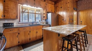 Eine Küche mit Holzausstattung, einer Granitarbeitsplatte, einem Edelstahlkühlschrank und einem Fenster mit Jalousien.