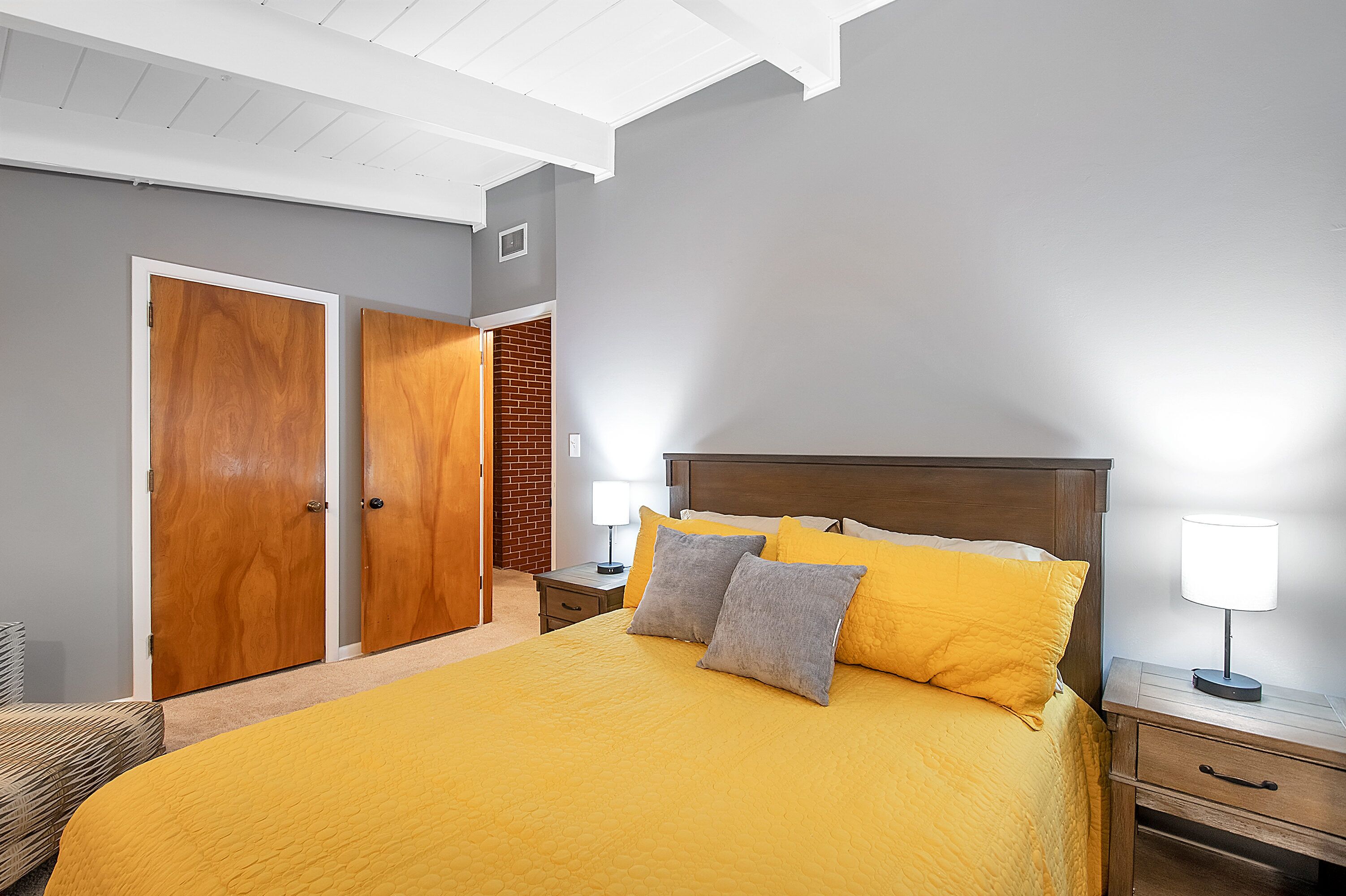 A bedroom with a large bed, two wooden wardrobes, a nightstand with a lamp, and a brick wall.