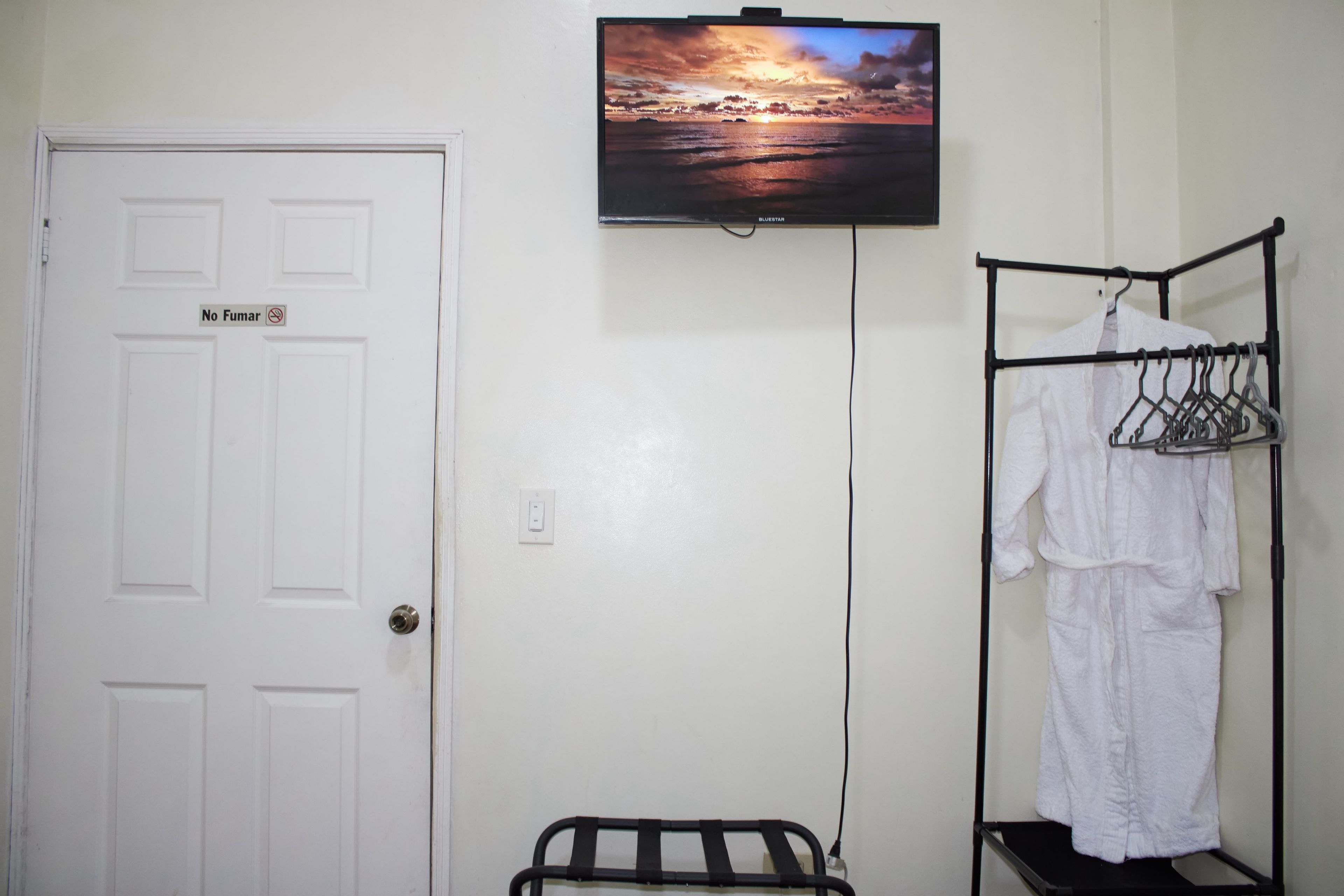 A room with a white door, a television mounted on the wall displaying a sunset scene, a clothing rack with a white robe, and a small black shelf.