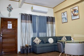 Uma sala de estar com sofá, mesa pequena, cadeira e janela com cortinas.