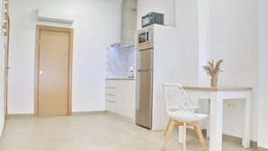 A modern kitchen with a built-in oven, microwave, and refrigerator. There is a wooden door and a white chair with a vase of dried flowers on a small table. - La Ilusión IV, studio in Conil. (Conil de la Fra.)