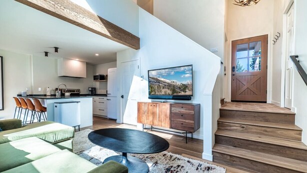 A modern living room with a sofa, a coffee table, and a TV on a wooden cabinet. There is a staircase leading to a door with a glass panel. - Sweet 1Bed/Loft/1Bath EagleVail (Avon)