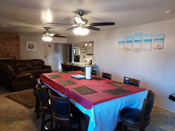A dining area with a long table covered in a red tablecloth, surrounded by chairs. There are ceiling fans above the table, and a kitchen area is visible in the background.