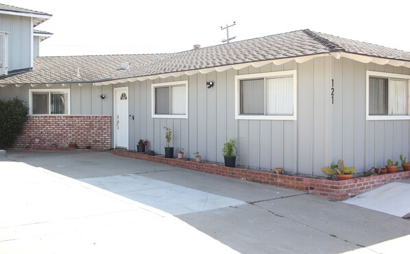 A single-story house with a grey exterior, a tiled roof, and a brick foundation. There are four windows, a front door, and a small porch area with potted plants. - Cozy home near the beach (Grover Beach)
