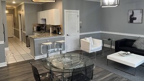 A modern kitchen with white cabinets, a dining area with a glass table, and a living room with a black sofa and a white coffee table. - Relaxing Now and Zen: Modern Condo, Near UNCC, PNC Pavilion, Pool, 5 Min to Dining, Shops, +15% off! (Charlotte)