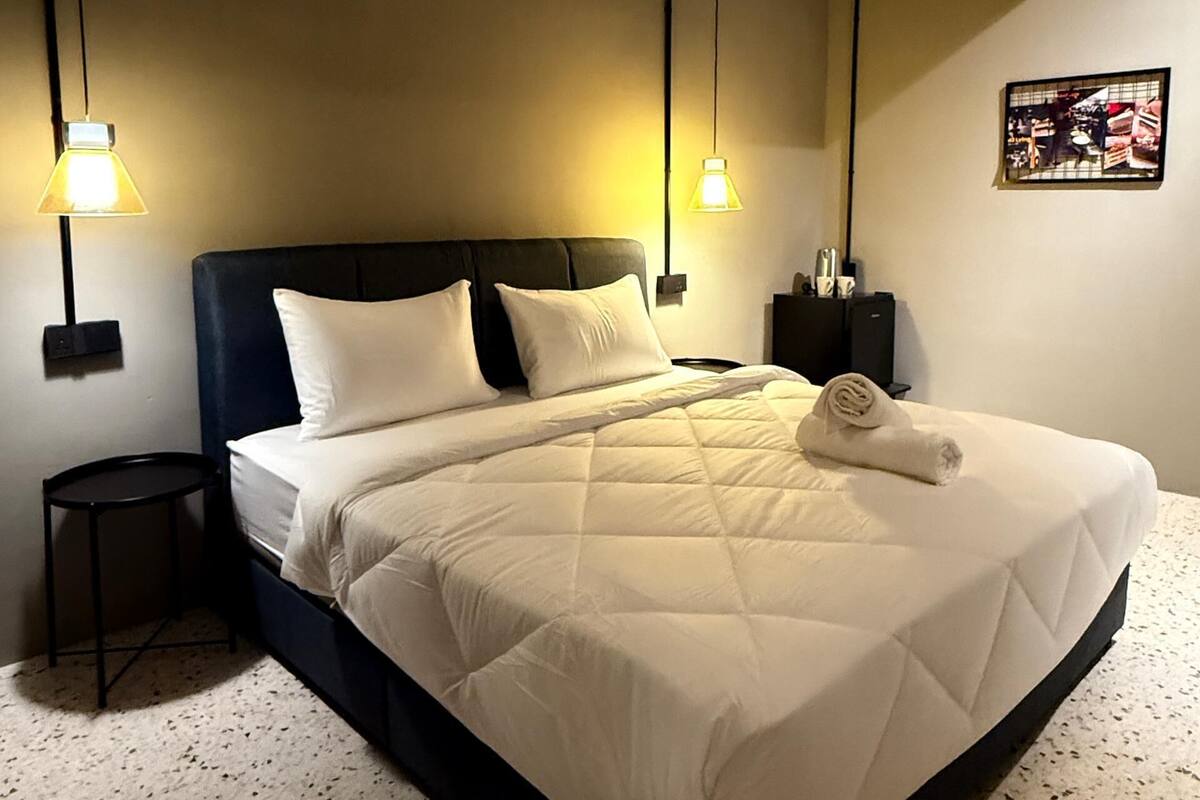 A neatly made bed with a quilted white duvet, two bedside lamps, a small round table, and a framed picture on the wall.