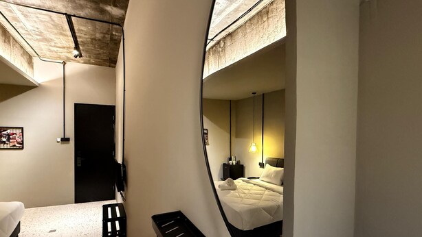 Una habitación de hotel moderna con un espejo grande que refleja la cama y una mesita de noche. La habitación tiene techo de concreto y un escritorio pequeño con una silla.