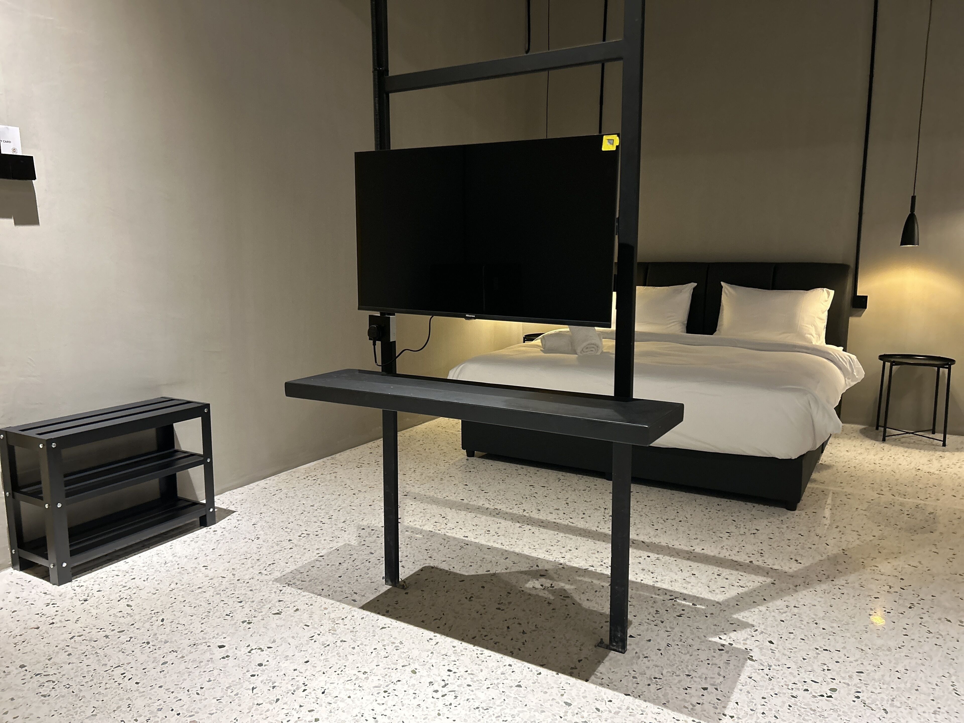 A modern hotel room with a bed, a TV mounted on a black stand, a small side table, and a shelf unit.