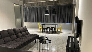 Una sala moderna con un sofá oscuro, una mesita redonda, una mesa de comedor con sillas y un televisor montado en la pared.