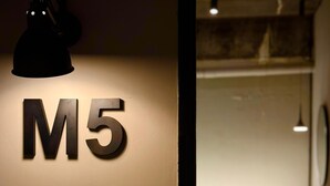 Una pared con el número 'M5' iluminado, una lámpara colgante y una vista parcial del interior de un baño.