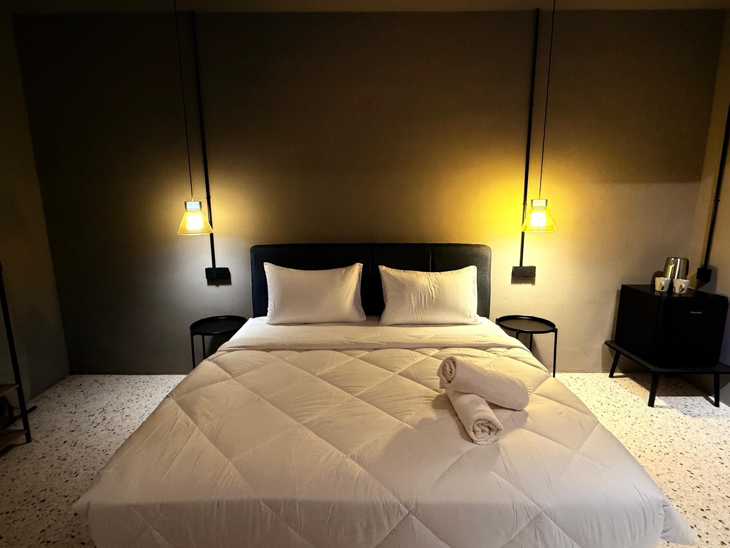 Un lit bien fait, recouvert d’une couverture matelassée, avec deux tables de chevet et des lampes fixées au mur.