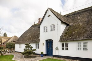 Een klassiek wit huis met rieten dak, een blauwe deur en een pad van stenen dat naar de ingang leidt.