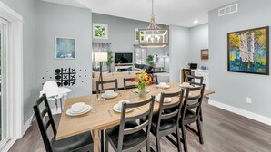 Un comedor moderno con una mesa de madera dispuesta para seis personas, rodeada de sillas oscuras. Un candelabro cuelga del techo, y un cuadro decora la pared.