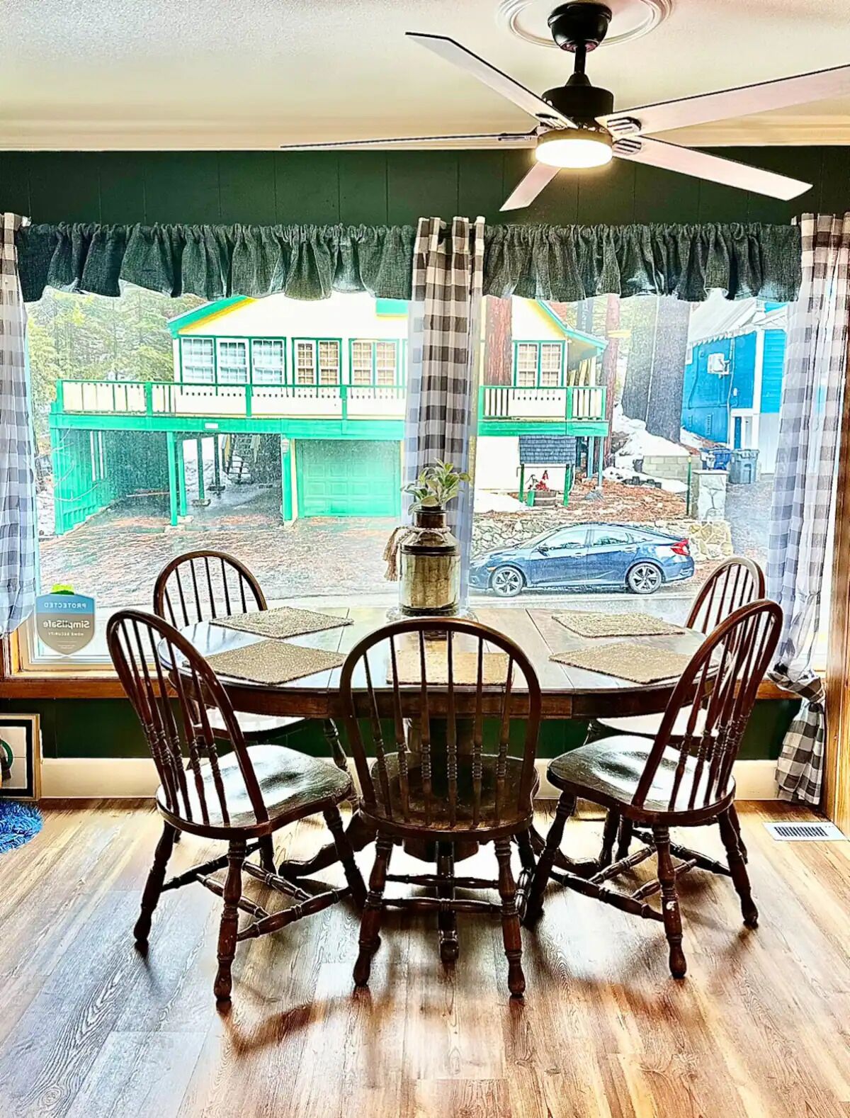 Un coin repas comprenant une table et quatre chaises, un ventilateur de plafond et une vue sur une rue bordée d’immeubles et de voitures garées.