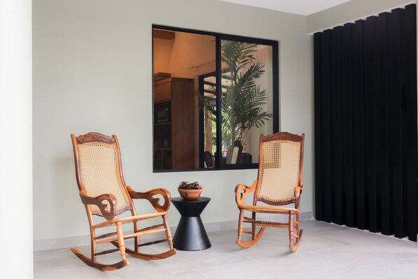 Une pièce avec deux fauteuils à bascule, une petite table sur laquelle est posée une plante en pot, et une grande fenêtre donnant sur un jardin.