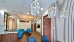 A modern living room with a wooden floor, a blue sofa, a dining table, and two crystal chandeliers. - Asahi Luxstay - Golden Park  Apartments (Hanoi)