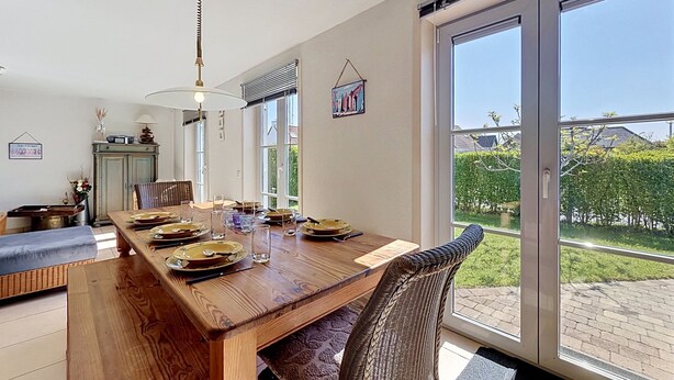 A dining area with a wooden table set for four, a wicker chair, and a view of a garden through large sliding glass doors.