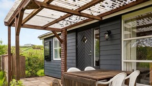Una pérgola de madera con una mesa y sillas, una pared exterior oscura y una ventana con vistas al entorno verde.