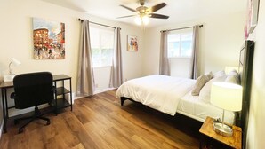 A bedroom with a bed, a desk, a chair, a ceiling fan, and artwork on the walls.