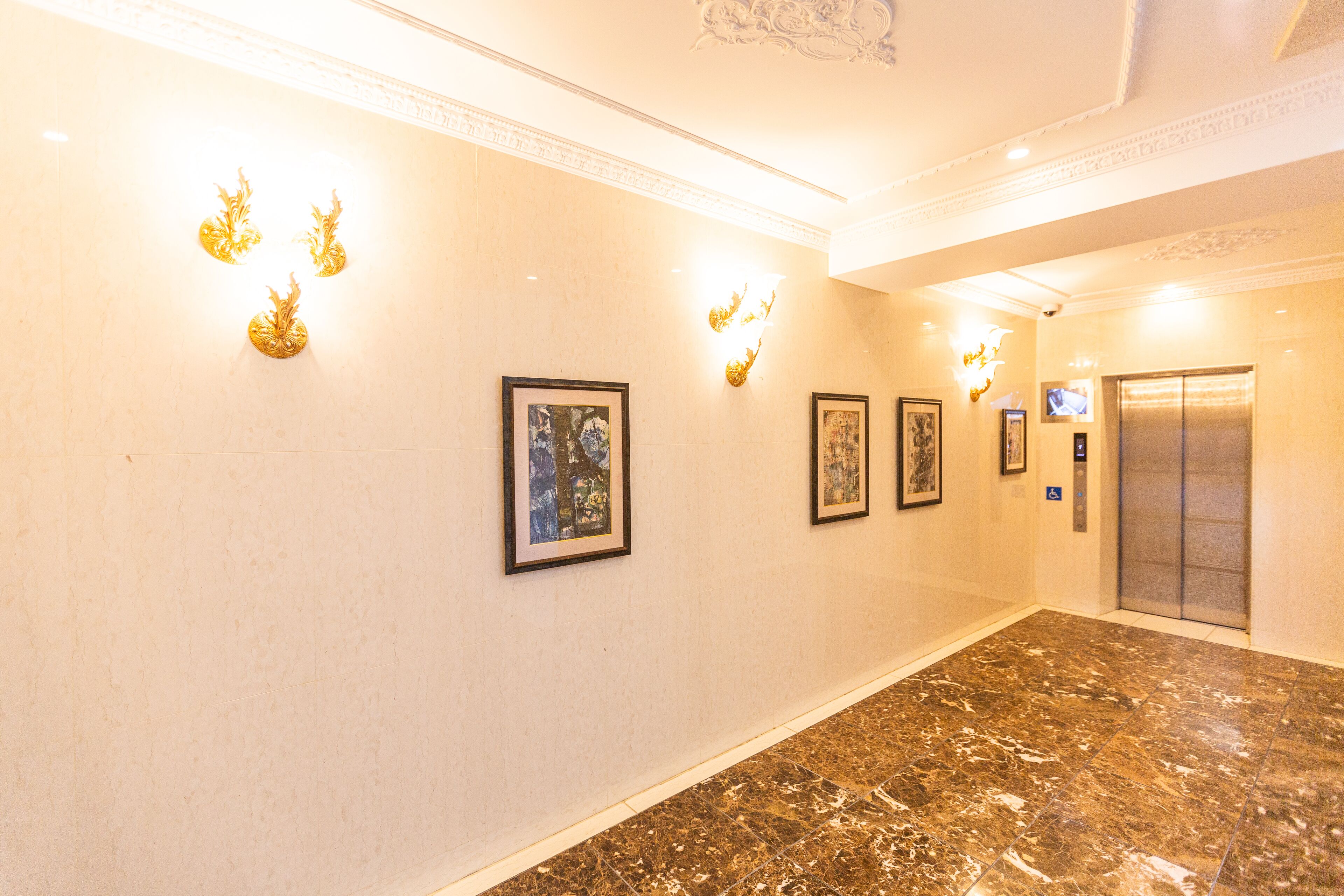 A hallway with marble flooring, wall-mounted light fixtures, framed artwork, and an elevator.