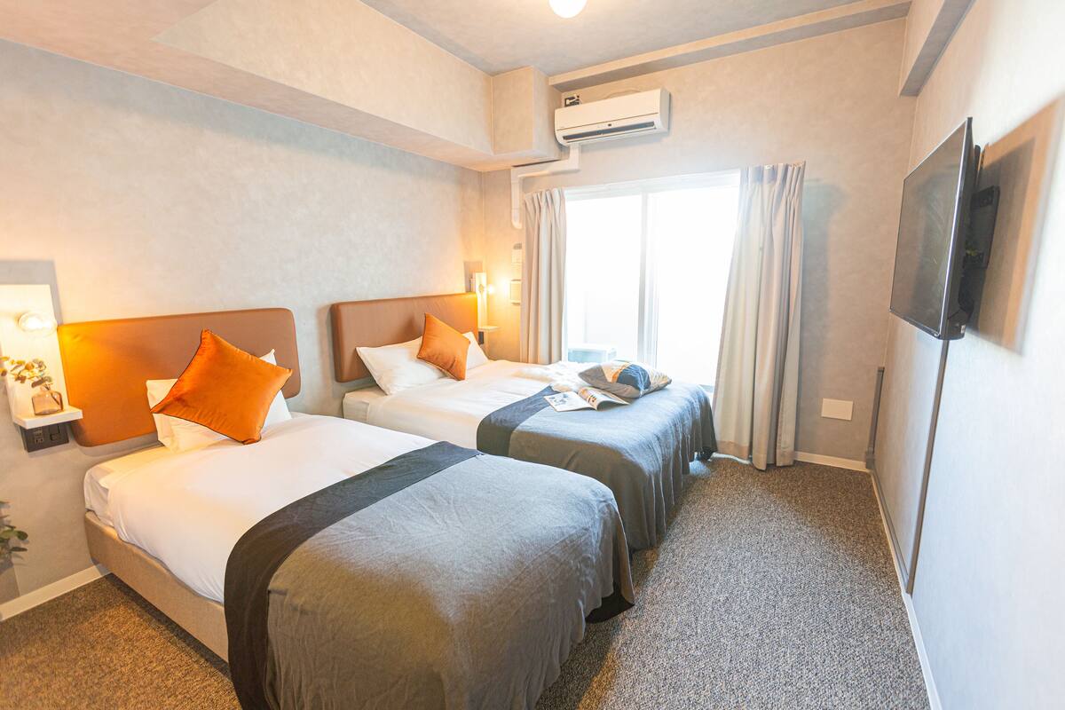 Una habitación de hotel con dos camas, un televisor de pantalla plana, un equipo de aire acondicionado y una ventana con cortinas.