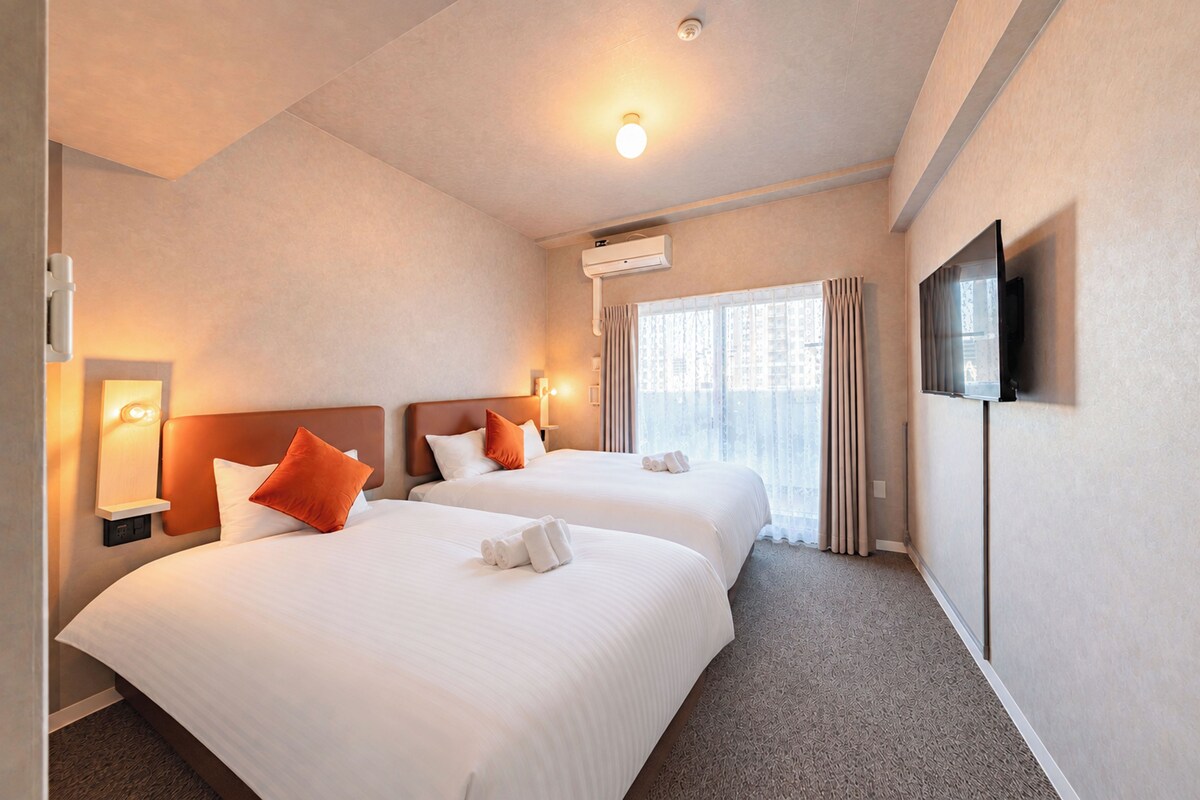 A hotel room with two beds, a flat-screen TV, an air conditioning unit, and a window with curtains.