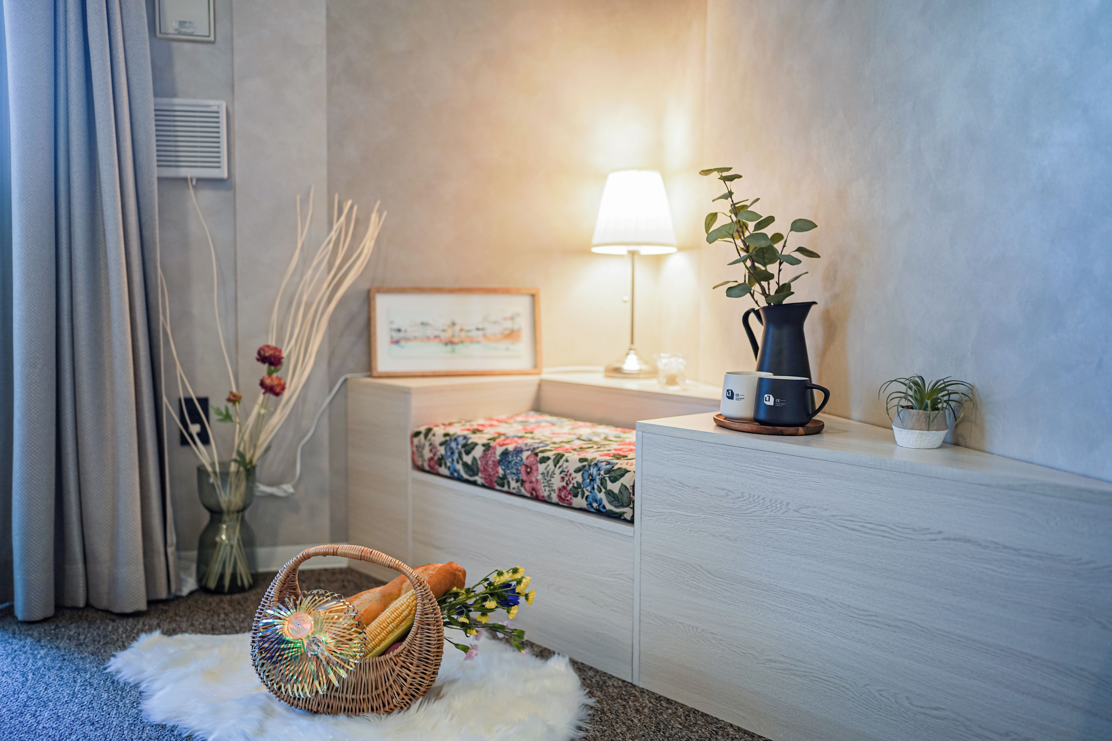 A neatly arranged hotel room with a bed, a bedside table with a lamp, a small potted plant, and a wicker basket on the floor.