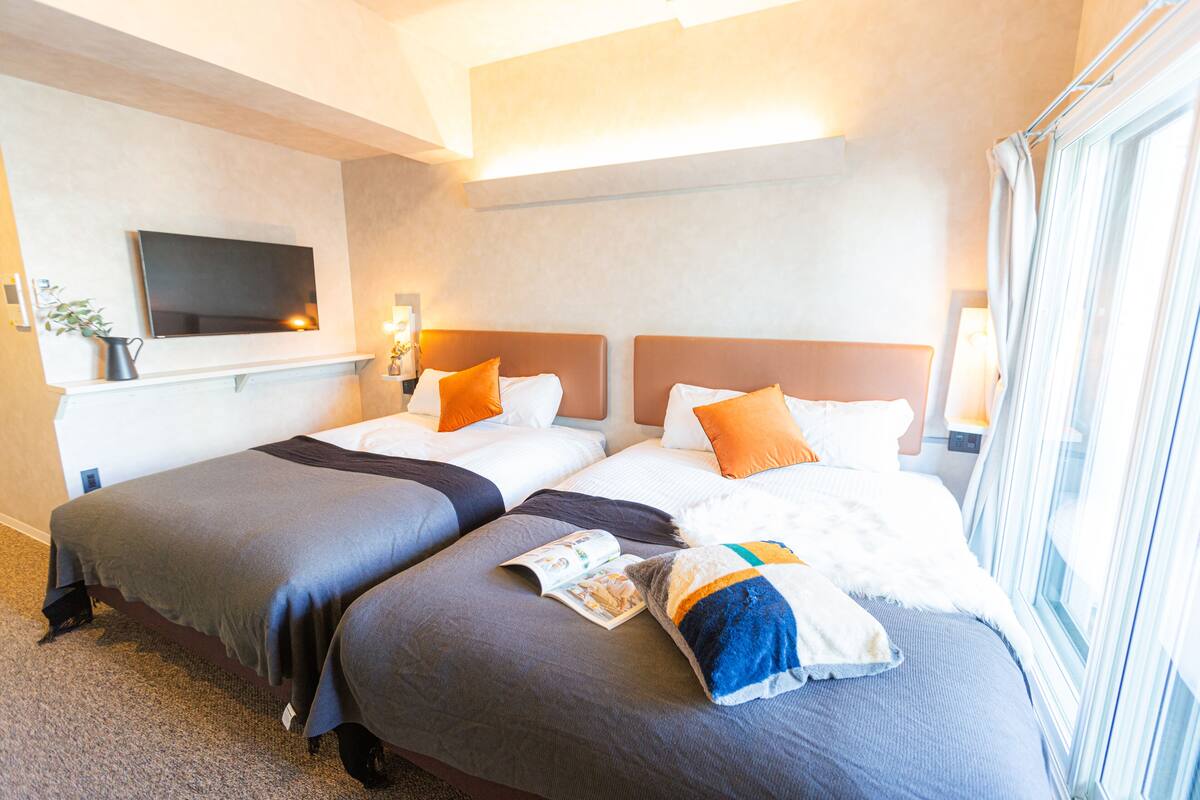 Habitación de hotel con dos camas, un televisor de pantalla plana montado en la pared y una ventana con cortinas.