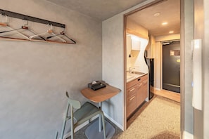 A compact hotel room with a small desk, a chair, a wardrobe, and a kitchenette.