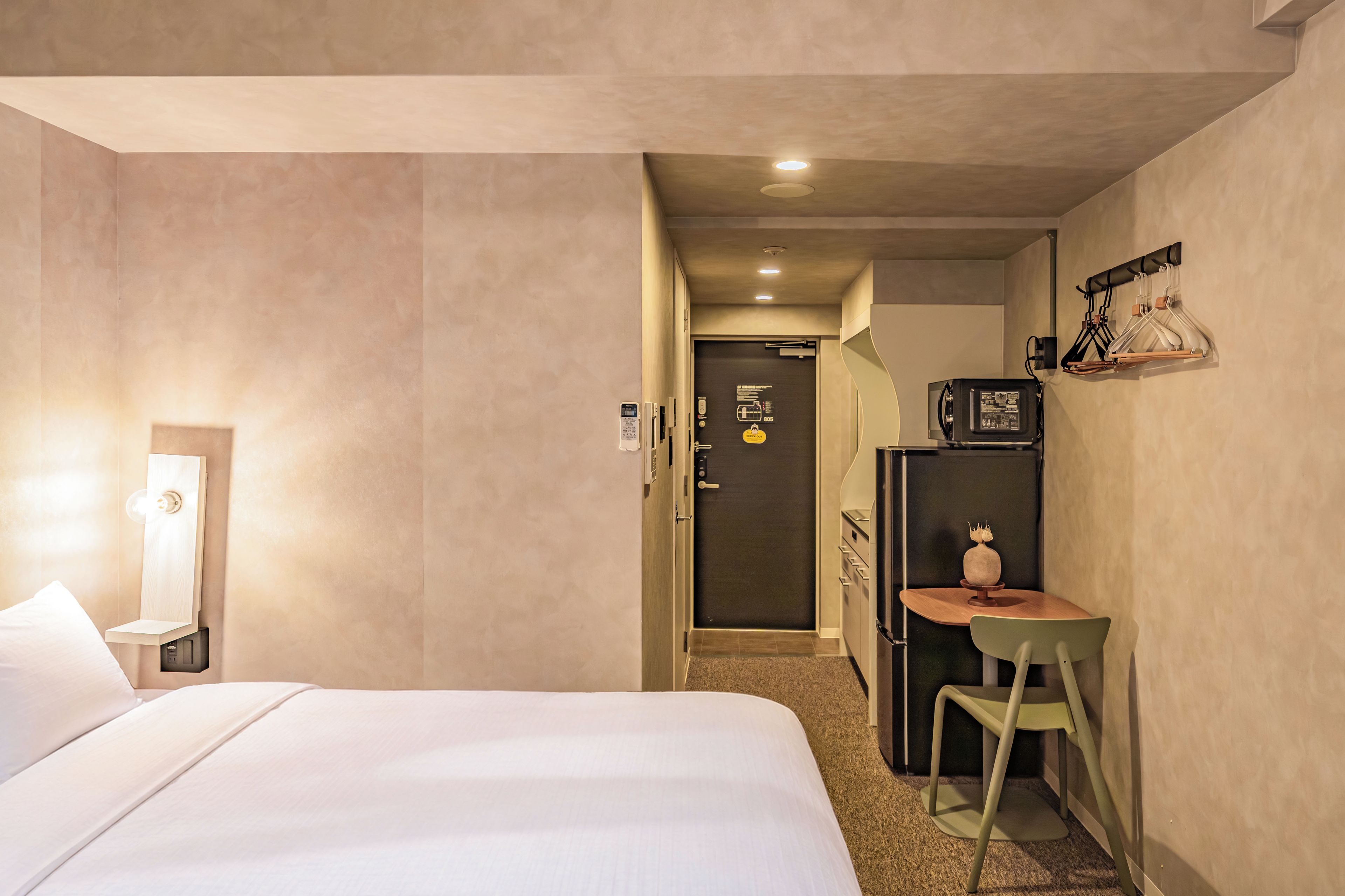 A hotel room with a bed, a desk, a chair, a small table, and a refrigerator.