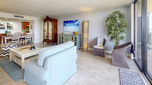 A spacious living area with a sofa, chairs, a dining table, and a large window. - Island Living! Beachfront Condo. Local Art. Heated Pool, Tennis, Spacious, Private & Secure. (Treasure Island)