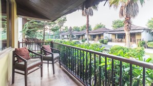 A balcony with wooden chairs and a railing, overlooking a courtyard with palm trees and thatched-roof buildings. - Treebo Tiger Pug Mark Resort with Pool (Kaladhungi)