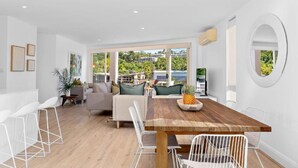 A spacious living area with a wooden dining table, white chairs, and a sofa with green cushions. - Edgewater Collection by Stay Noosa (Noosa Heads)