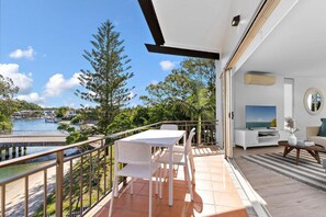 A balcony with a white table and chairs, a TV, and a view of a body of water and greenery. - Edgewater Collection by Stay Noosa (Noosa Heads)