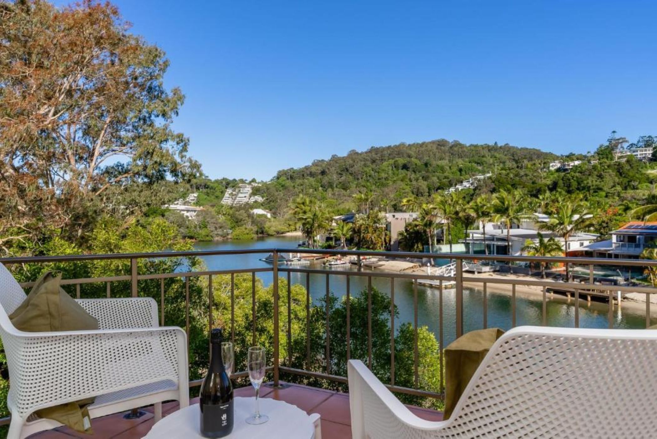 Un balcon avec des fauteuils en osier, une table avec une bouteille et un verre, donnant sur une rivière où l’on aperçoit des bateaux et des maisons au loin.