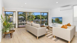 A modern living room with a sofa, a coffee table, and a TV. There is a large window with a view of the outdoors. - Edgewater Collection by Stay Noosa (Noosa Heads)