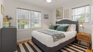 A bedroom with a bed, bedside lamps, a ceiling fan, and large windows with blinds. - Sail Away - Pet Friendly! (Jekyll Island)