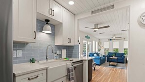 A modern kitchen with white cabinets, a blue backsplash, and a wooden floor leading to a living area with blue sofas and a coffee table. - Sail Away - Pet Friendly! (Jekyll Island)