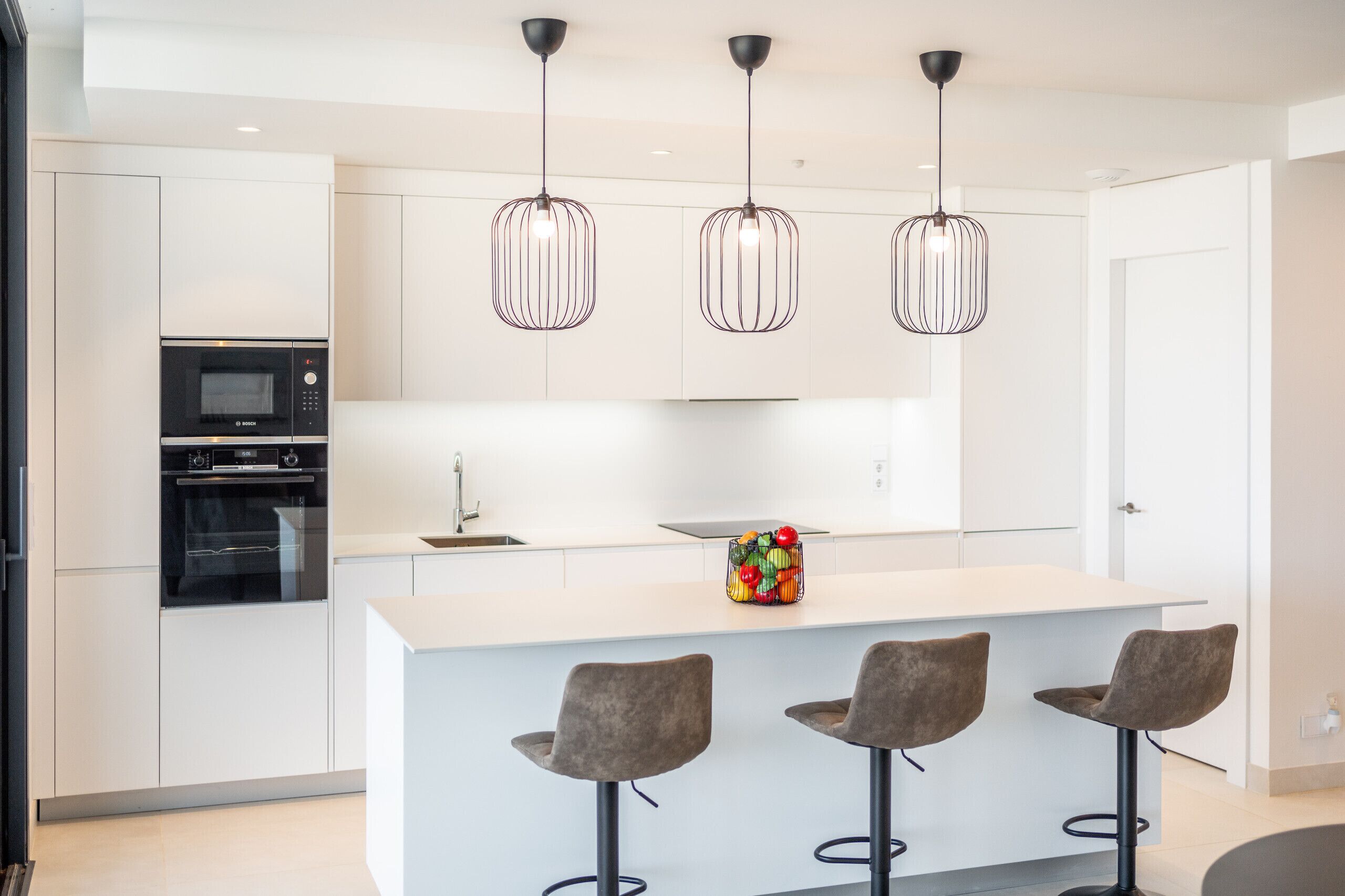 A modern kitchen with white cabinets, a built-in oven, a large island with bar stools, and pendant lighting.