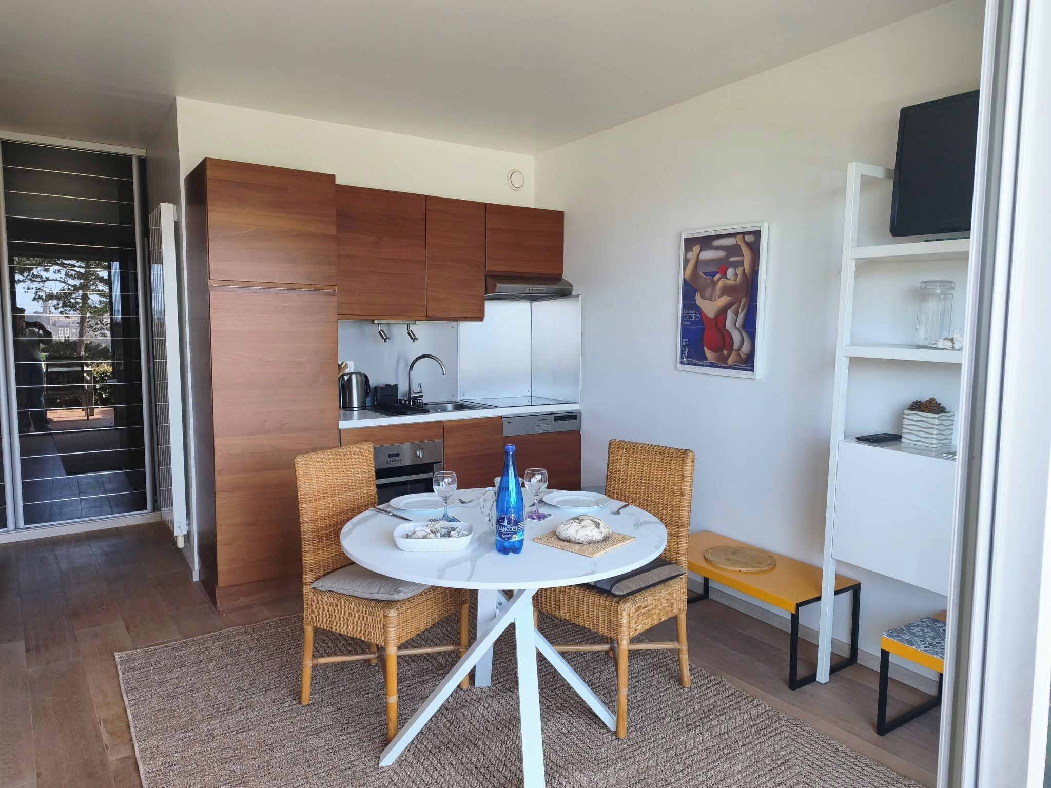 A modern kitchen with wooden cabinets, a dining area with wicker chairs, and a small white table with a glass of water.