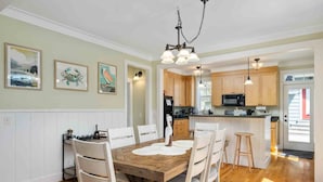 A dining area with a wooden table and white chairs, a kitchen with wooden cabinets, and a hanging light fixture.