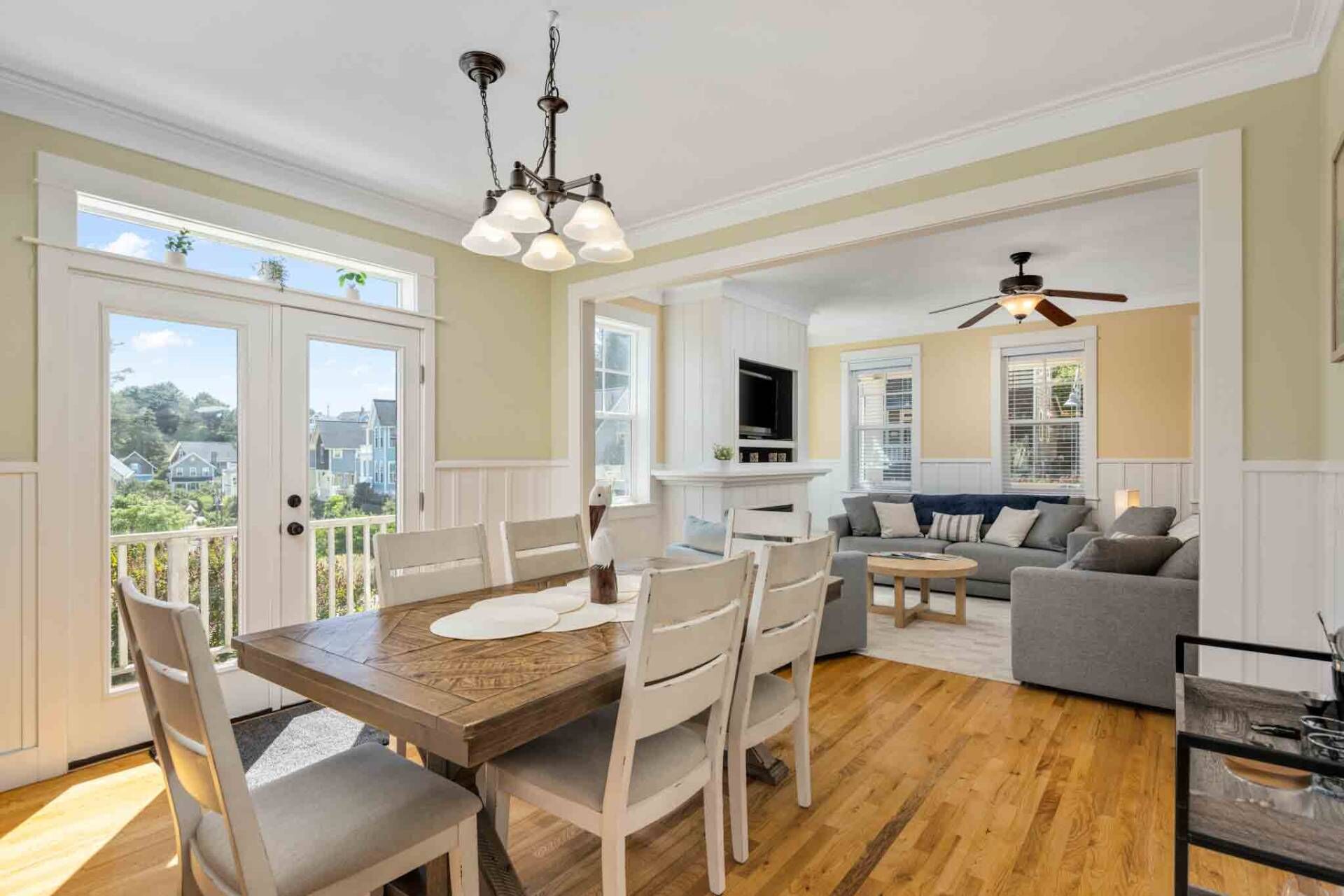 Un coin repas avec une table et des chaises en bois, un ventilateur de plafond et une vue sur le quartier grâce à de grandes fenêtres.