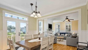 A dining area with a wooden table and chairs, a ceiling fan, and a view of the neighborhood through large windows.