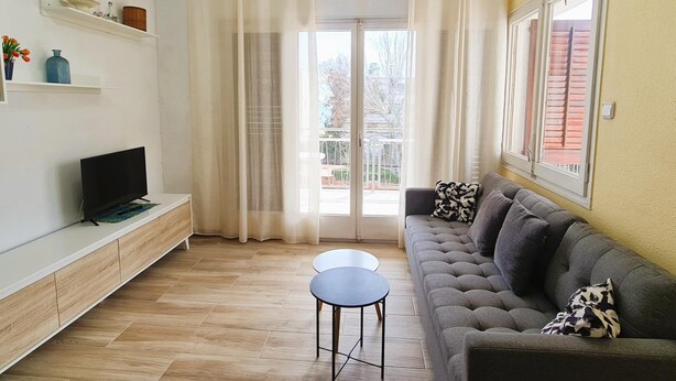 Un salon avec un canapé, une petite table, un téléviseur posé sur un meuble, une porte-fenêtre donnant sur un balcon et une fenêtre avec des rideaux.