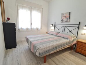 A bedroom with a bed, a nightstand, a dresser, a window with curtains, and a wall-mounted light fixture.