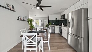 Cozinha moderna com armários brancos, uma geladeira de aço inoxidável e uma área de jantar com mesa de madeira e cadeiras brancas.