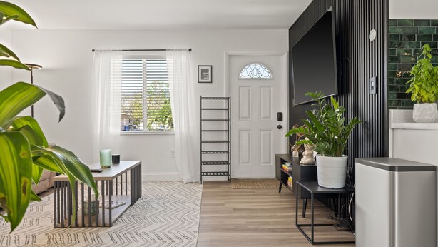 Uma sala de estar moderna com piso de madeira, um tapete estampado, uma mesa de metal preto, uma porta branca e uma planta verde.
