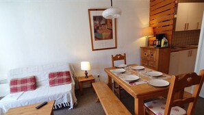 A cozy living room with a sofa, a wooden dining table set for four, a bench, and a kitchen area with cabinets and a hanging light fixture. - Cosy apt 4/6 ppl with balcony, near the ski runs (Les Deux Alpes)