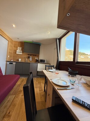 Een compacte keuken met houten kastjes, een eettafel gedekt voor twee personen, en uitzicht op een besneeerd landschap door een raam.